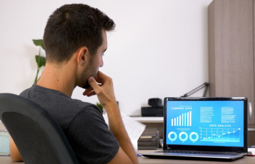 A person is sitting at a desk, looking at a laptop screen. The laptop displays a presentation with various charts and graphs, including a bar graph, pie chart, and line graph. The person appears to be deep in thought, with one hand on their chin. The room has a minimalistic design with a plant in the background and a lamp on the desk.