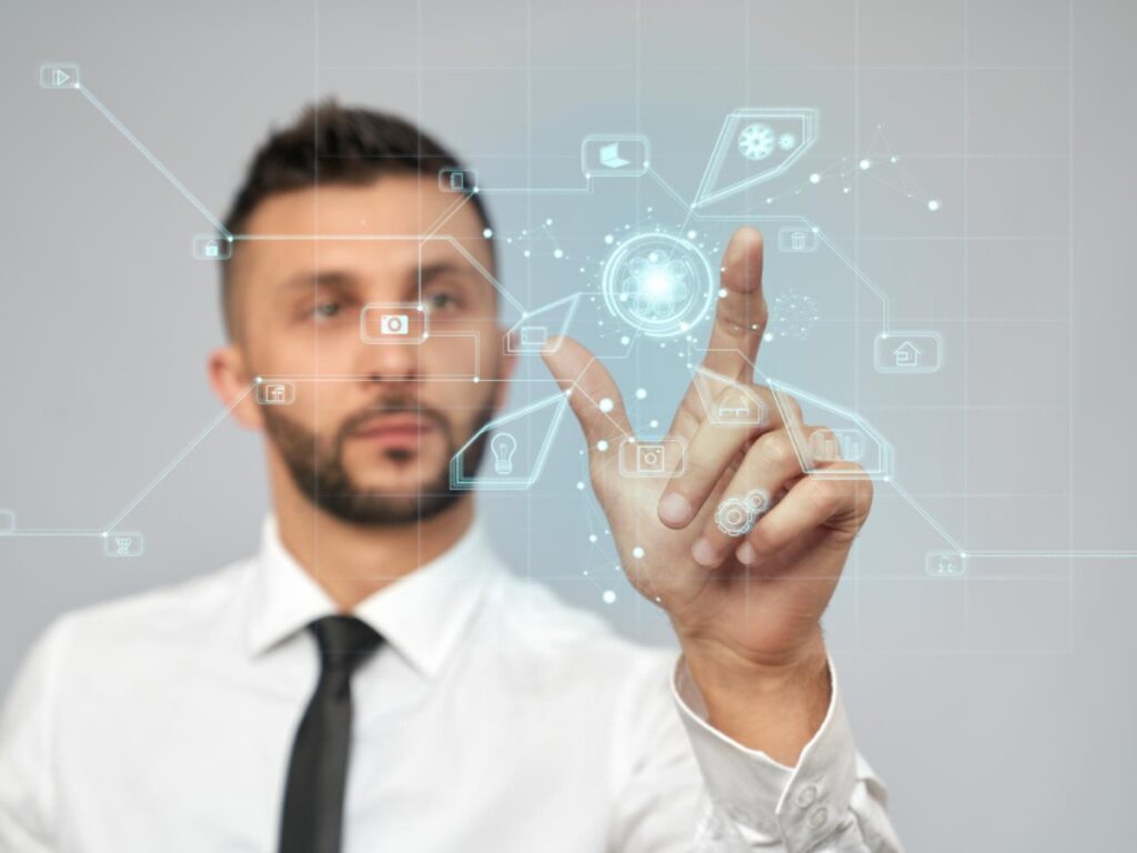 How to describe your business with AI, the image shows a person in a white shirt and black tie, pointing at a digital interface. The interface is composed of various icons and lines, suggesting a futuristic or technological theme. The icons include symbols like a lock, a gear, a camera, and a speech bubble, which typically represent security, settings, photography, and communication, respectively. The background is a gradient gray, enhancing the high-tech appearance of the scene.