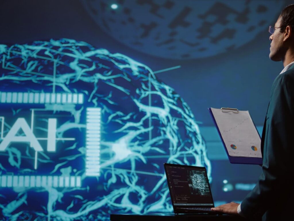 How to describe your business with ai, the image depicts a person in a professional setting, possibly a presentation or a meeting. The individual is holding a clipboard with a diagram or chart on it, and there is a laptop on a table in front of them. The background features a large screen displaying a brain-like structure with the word "AI" prominently displayed, suggesting a focus on artificial intelligence. The overall scene conveys a theme of technology and data analysis.