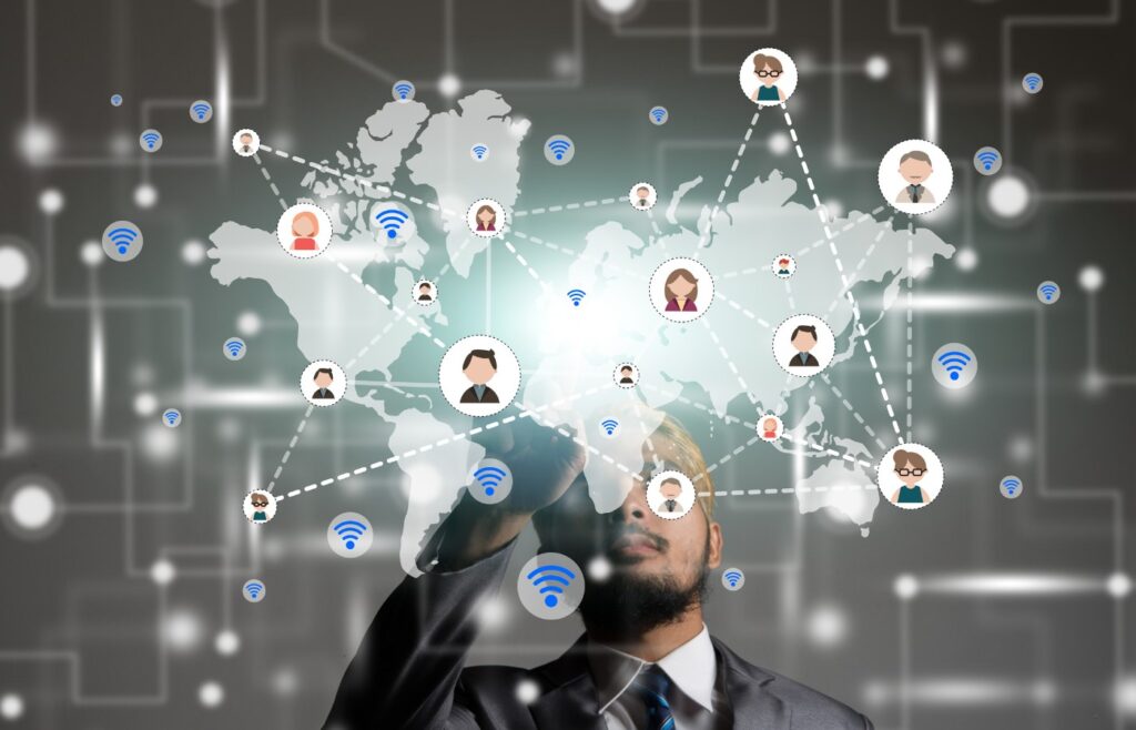 This image depicts a person in a suit looking at a digital representation of a global network. The background features a world map with various icons representing people and communication symbols like Wi-Fi and signal bars. The network is illustrated with dotted lines connecting these icons, symbolizing global connectivity and communication. The image conveys themes of international networking, digital communication, and interconnectedness.