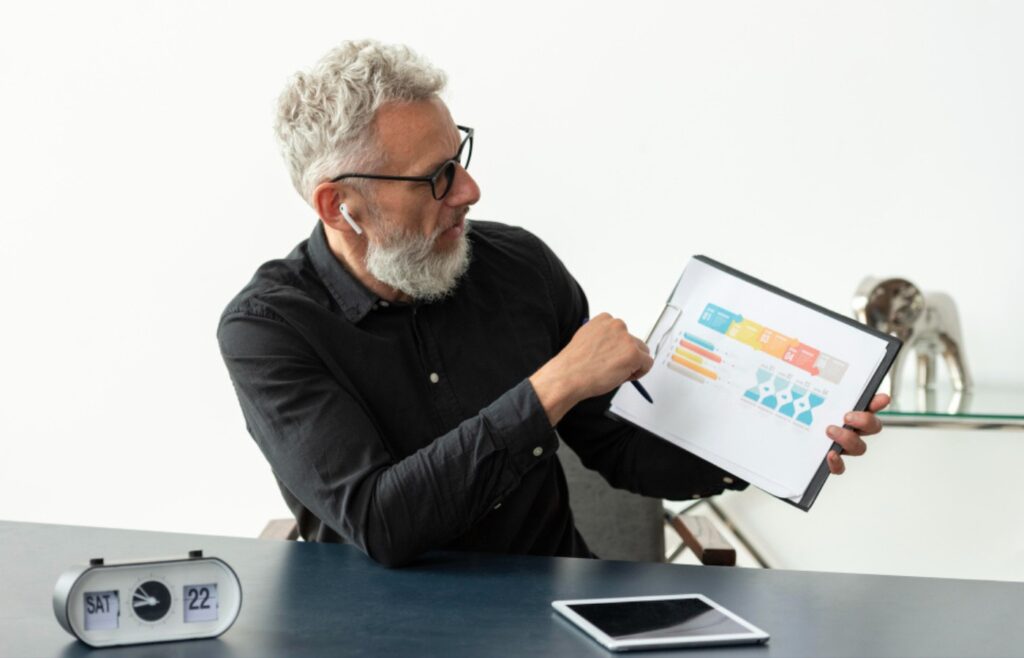 what does a digital marketing account manager do, The image shows a person sitting at a desk, holding a tablet displaying a colorful infographic. The person is wearing a black shirt and has earbuds in. On the desk, there is a digital clock showing the time as 22:17 and a tablet lying flat. The background includes a decorative elephant statue.